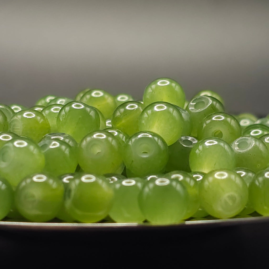 A close-up of glossy, translucent light green round beads with drilled holes, piled on a dark surface against a soft gray background.