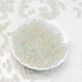 Small translucent pale green and white round beads with subtle gold flecks, displayed in a white scalloped dish with more beads scattered around on a light textured background.