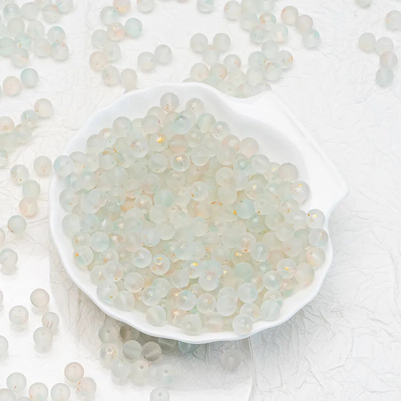 Small translucent pale green and white round beads with subtle gold flecks, displayed in a white scalloped dish with more beads scattered around on a light textured background.