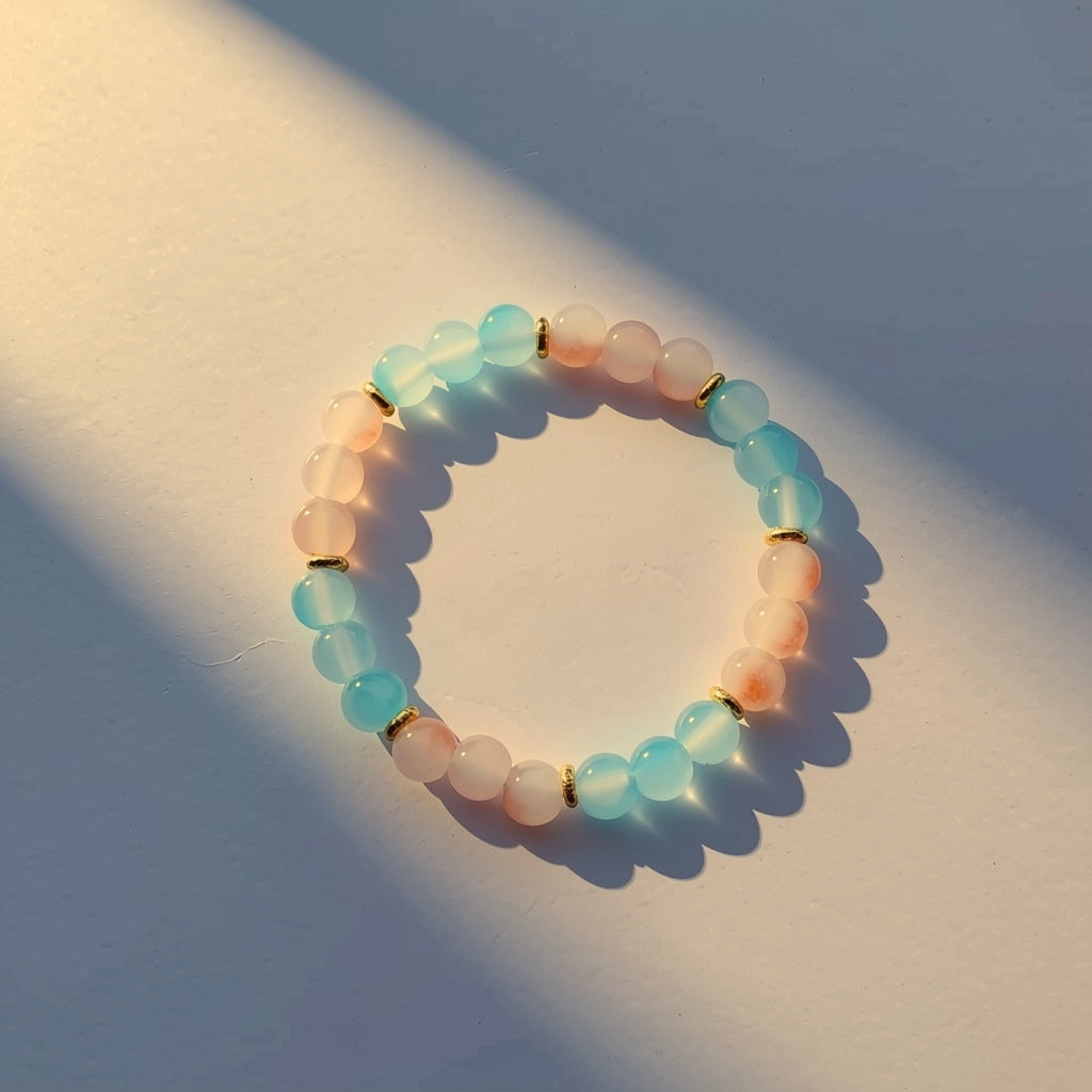 A beaded stretch bracelet made of translucent pastel blue and peach-pink round glass beads separated by small gold coloured spacer rings, photographed on a sunlit off-white surface with strong shadows.