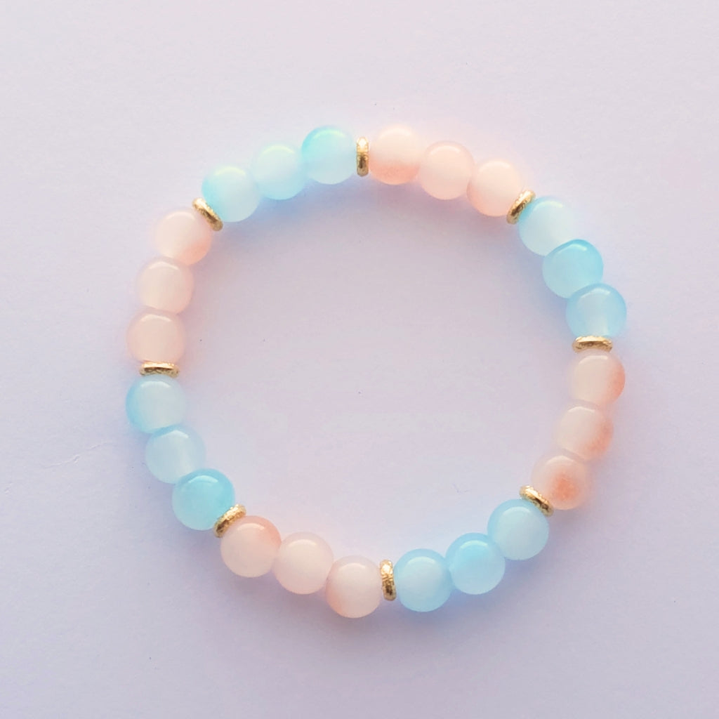A beaded stretch bracelet featuring alternating pastel light blue and peach-pink round glass beads, separated by small gold coloured spacer rings, shown on a light lavender background.