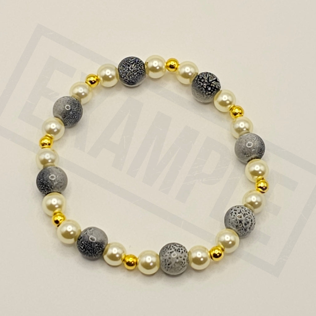 A beaded stretch bracelet featuring white faux pearls, gray marbled round beads, and small polished gold spacer beads, arranged in a circle on a light beige background.
