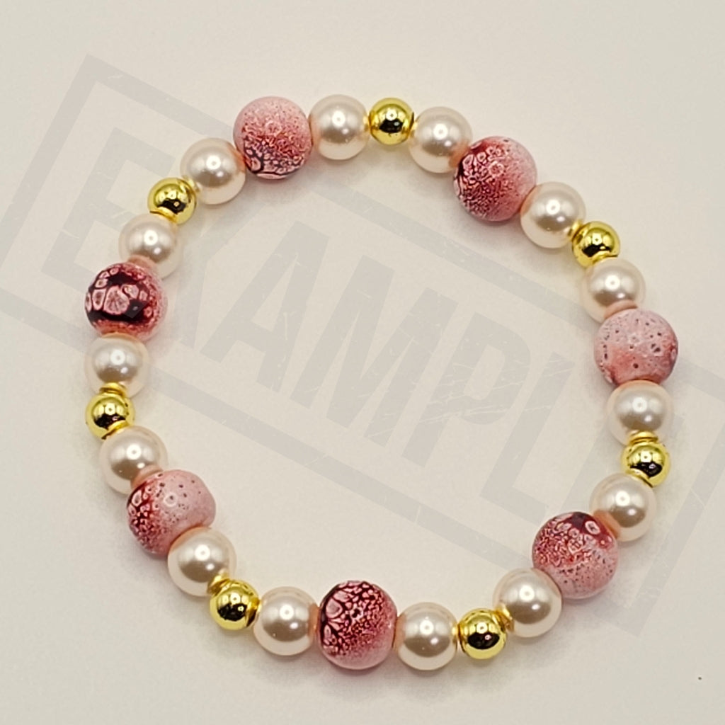 A beaded stretch bracelet featuring white faux pearls, pink marbled glass beads, and shiny gold spacer beads, arranged in a circle on a light background.