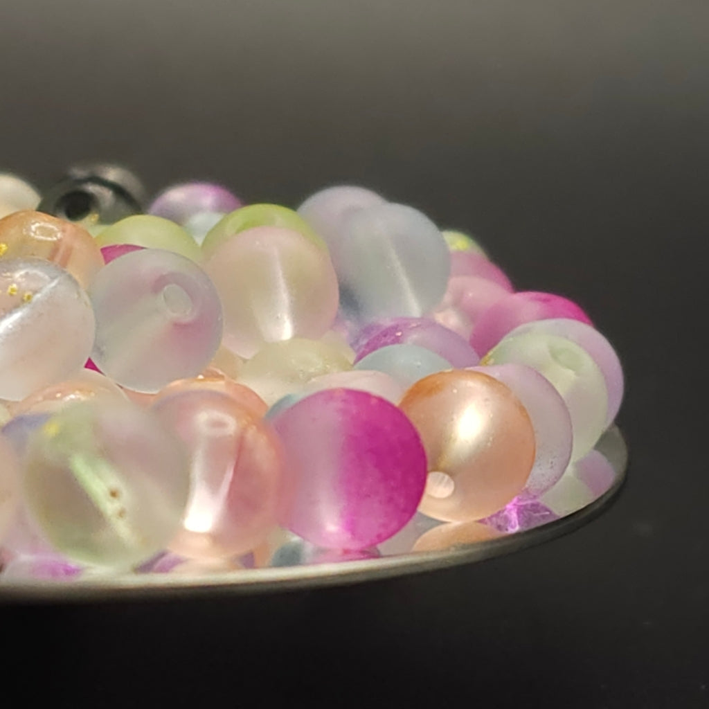 A close-up of translucent round beads in pastel colors including pink, peach, mint green, lavender, and pale yellow, arranged on a dark reflective surface.