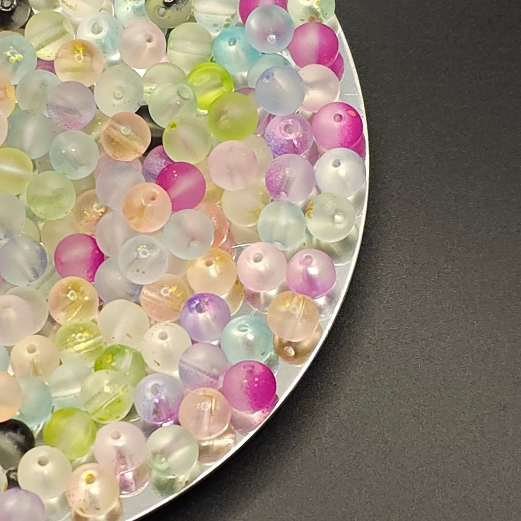 A close-up of many small translucent round beads in pastel colors, including pink, purple, blue, green, and clear tones, arranged on a silver tray against a dark gray background.