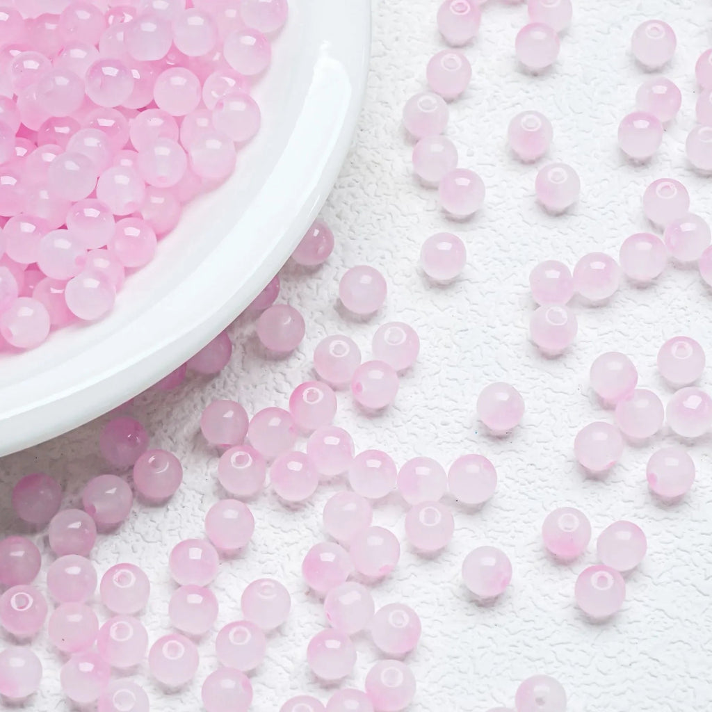 Pink translucent round beads scattered on a white textured surface, with a white bowl filled with more beads in the upper left corner.