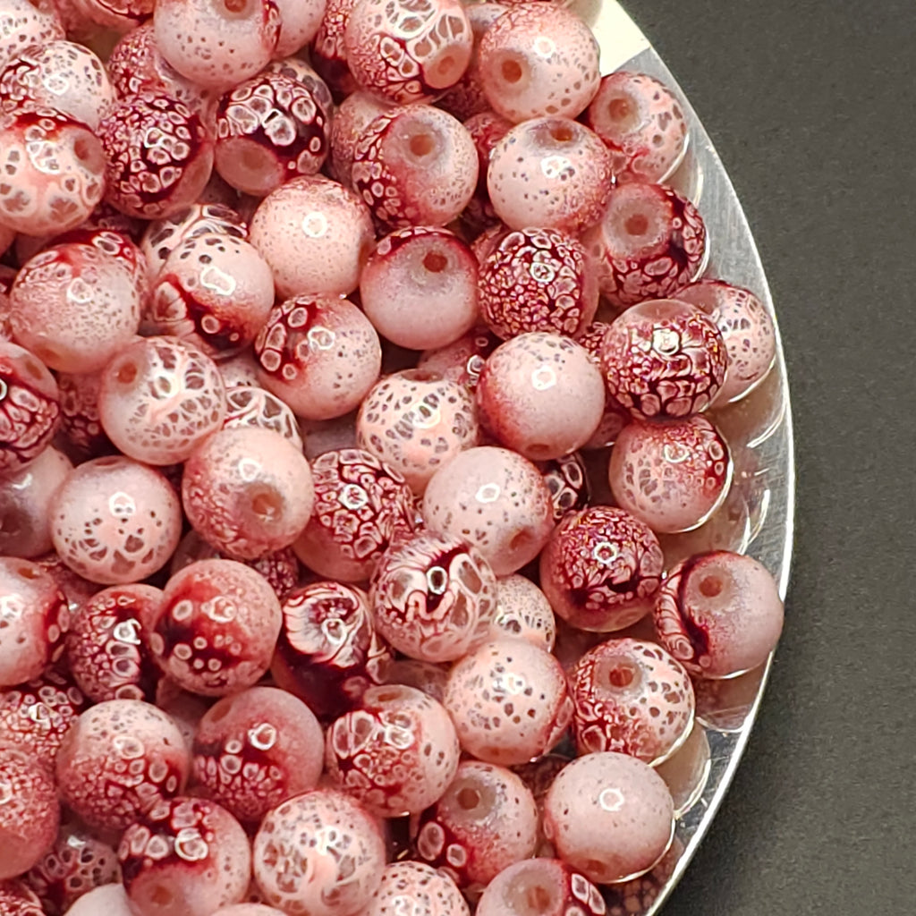 A close-up of many round glass beads in pale pink and translucent white with red mottled patterns, piled on a silver tray against a dark gray background.