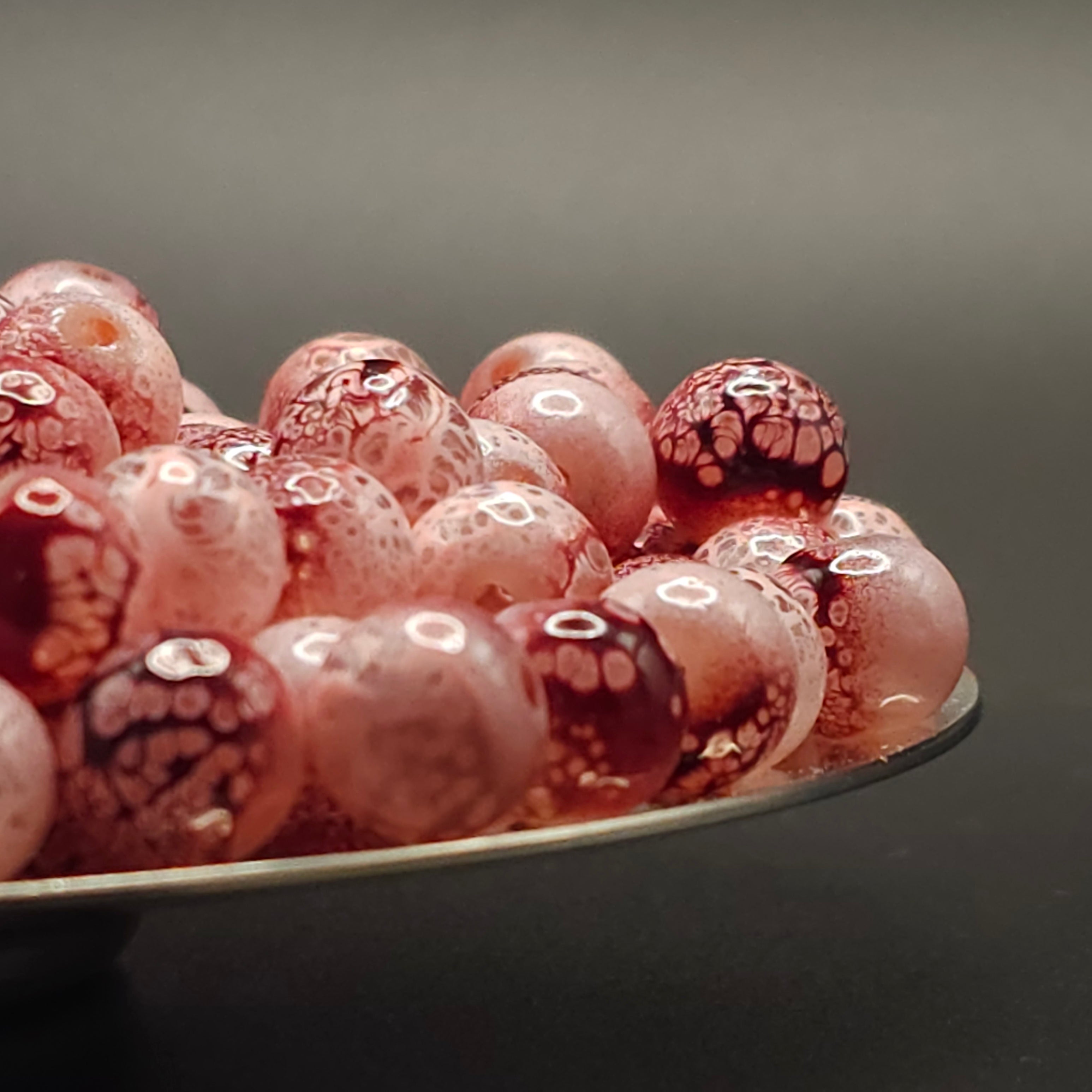 A close-up pile of round glass beads with a mottled pattern of deep red and pale pink, featuring white bubble-like speckles.