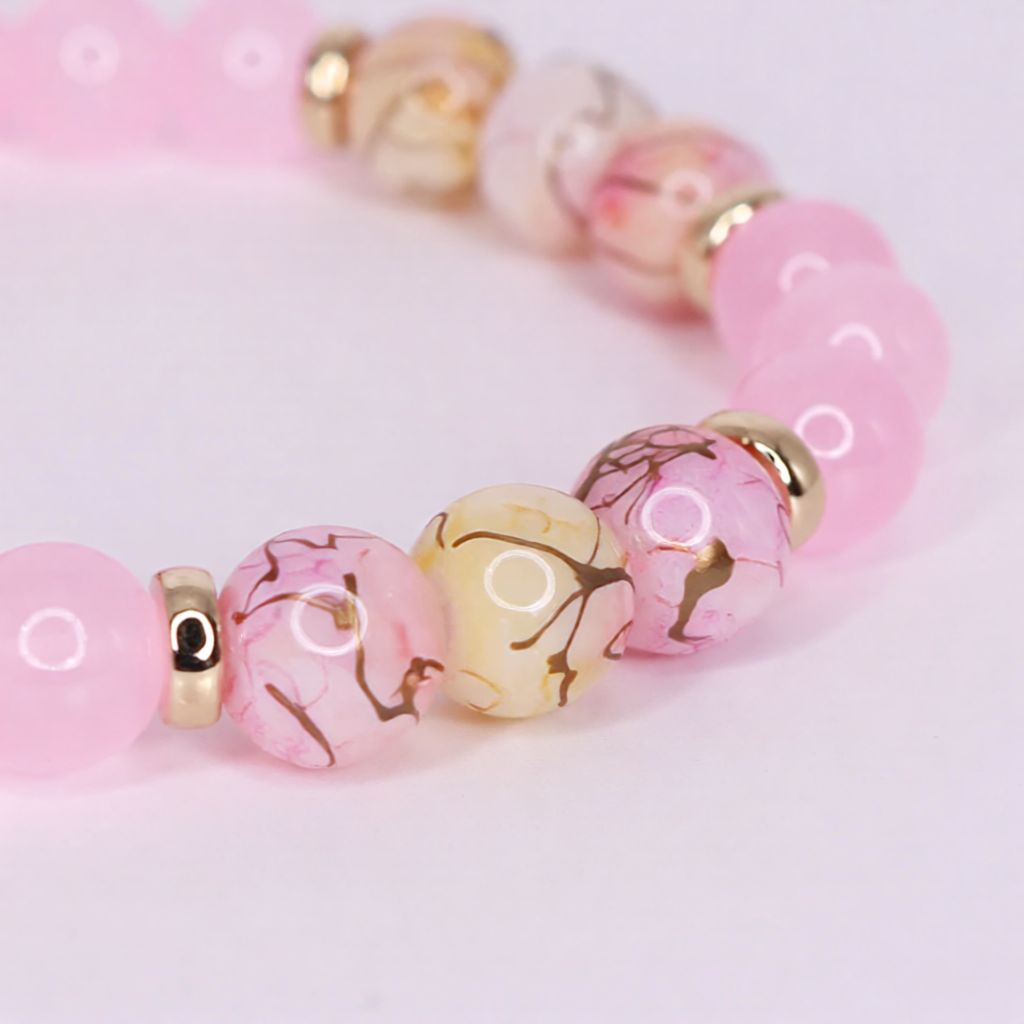 A close-up of a beaded glass bracelet featuring round pink beads, marbled pink and cream beads with gold branch-like patterns, and small gold coloured spacer beads, set against a soft light pink background.
