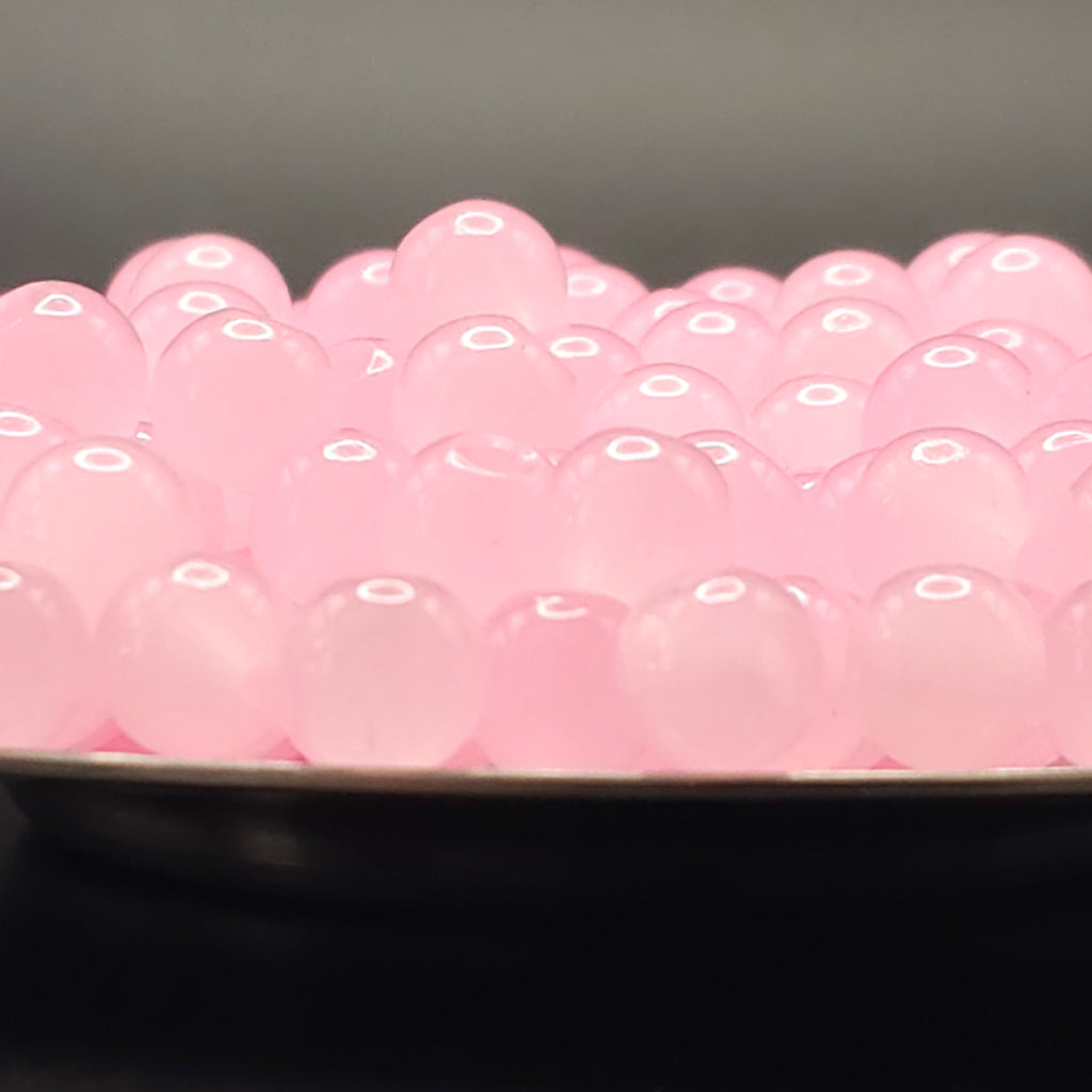 A close-up of many small translucent pink oval beads or boba pearls piled in a dark dish against a dark gray background.