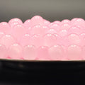 A close-up of many small translucent pink oval beads or boba pearls piled in a dark dish against a dark gray background.