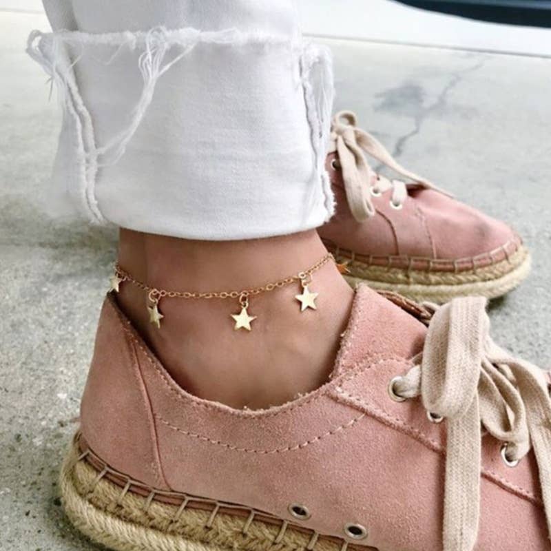 Pink espadrille shoes with a gold anklet featuring star charms on a light background.