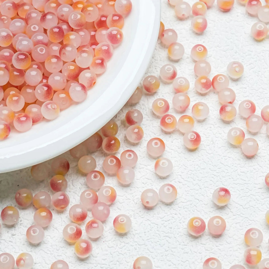 Pink translucent round beads with orange and yellow marbled patterns, shown in a white bowl with several scattered on a textured white surface.