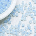 Light blue translucent round beads scattered on a white textured surface, with a white plate partially visible in the top left corner.