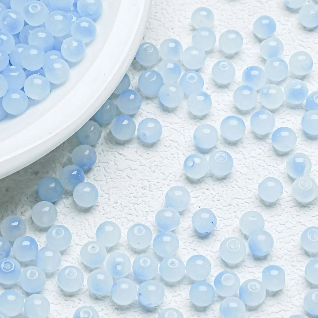 Light blue translucent round beads scattered on a white textured surface, with a white plate partially visible in the top left corner.