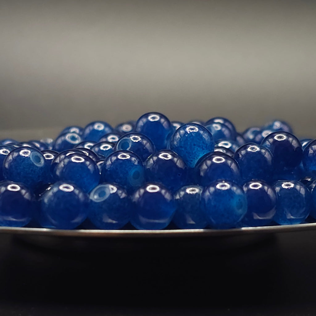 A pile of glossy, translucent deep blue glass beads resting on the edge of a dark plate against a dark gray background.