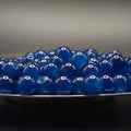 A pile of glossy, translucent deep blue glass beads resting on the edge of a dark plate against a dark gray background.