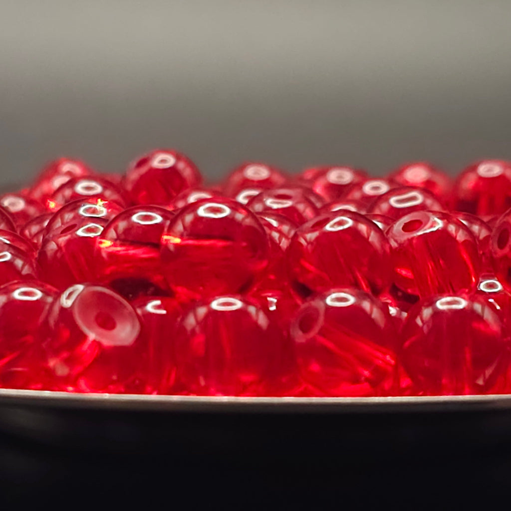 A close-up of many small translucent red round beads with holes, piled in a shallow dish against a dark gray background.
