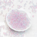 A white scalloped dish filled with small translucent round beads in pastel pink, lavender, and icy blue, with additional beads scattered around on a white textured background.