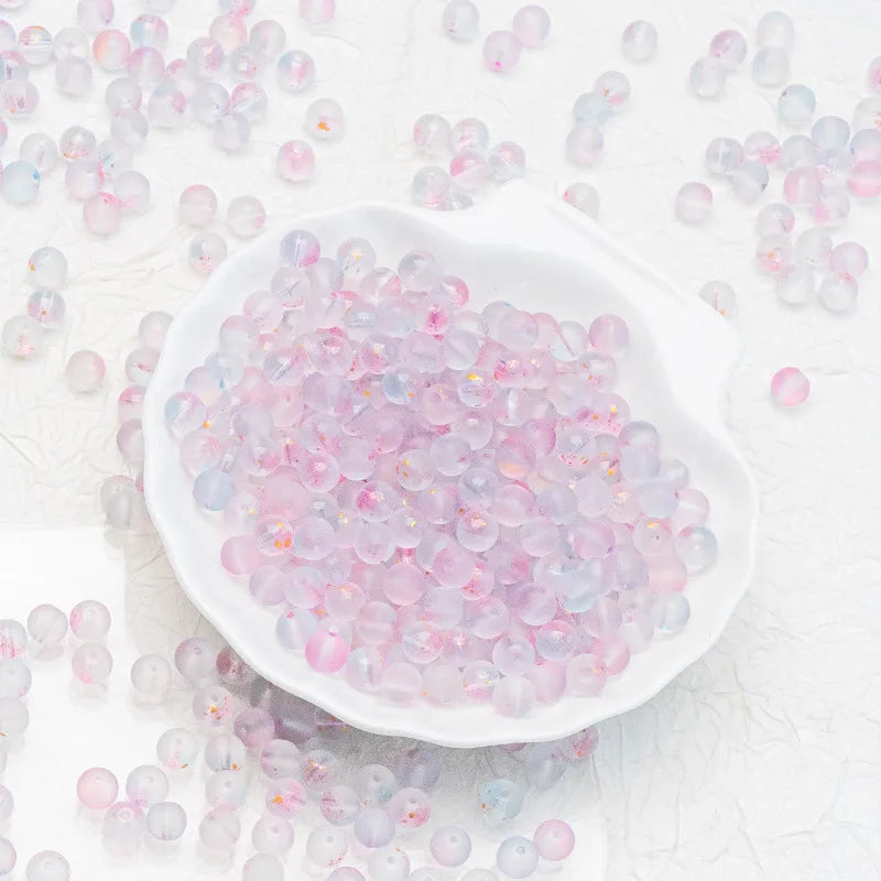 A white scalloped dish filled with small translucent round beads in pastel pink, lavender, and icy blue, with additional beads scattered around on a white textured background.