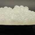 A close-up of many small translucent white oval beads or pearls piled in a dark dish against a dark gray background.