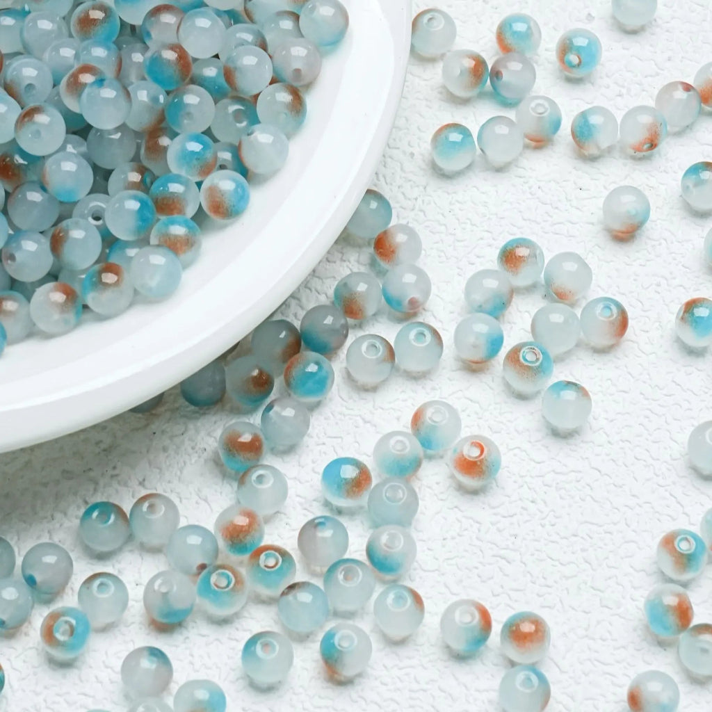Round marbled beads in shades of light blue, white, and orange-brown, with some beads spilled from a white plate onto a textured white surface.